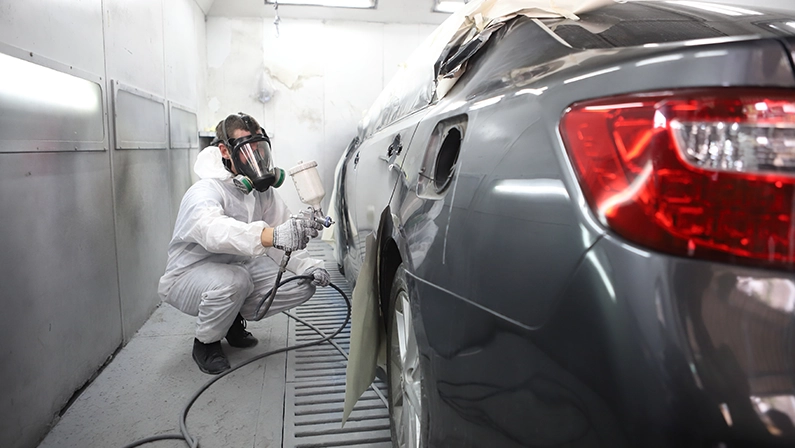 automobile repairman painter in protective workwear and respirator painting car body in paint chamber