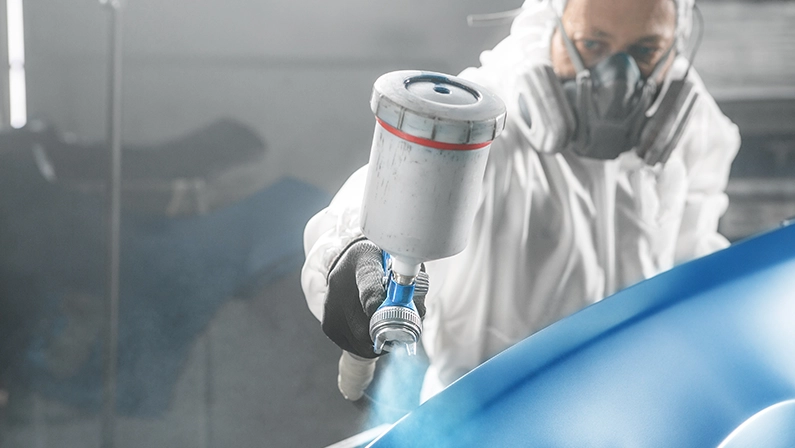 Car paint worker spraying blue paint to car body element using spray gun in vehicle workshop chamber.