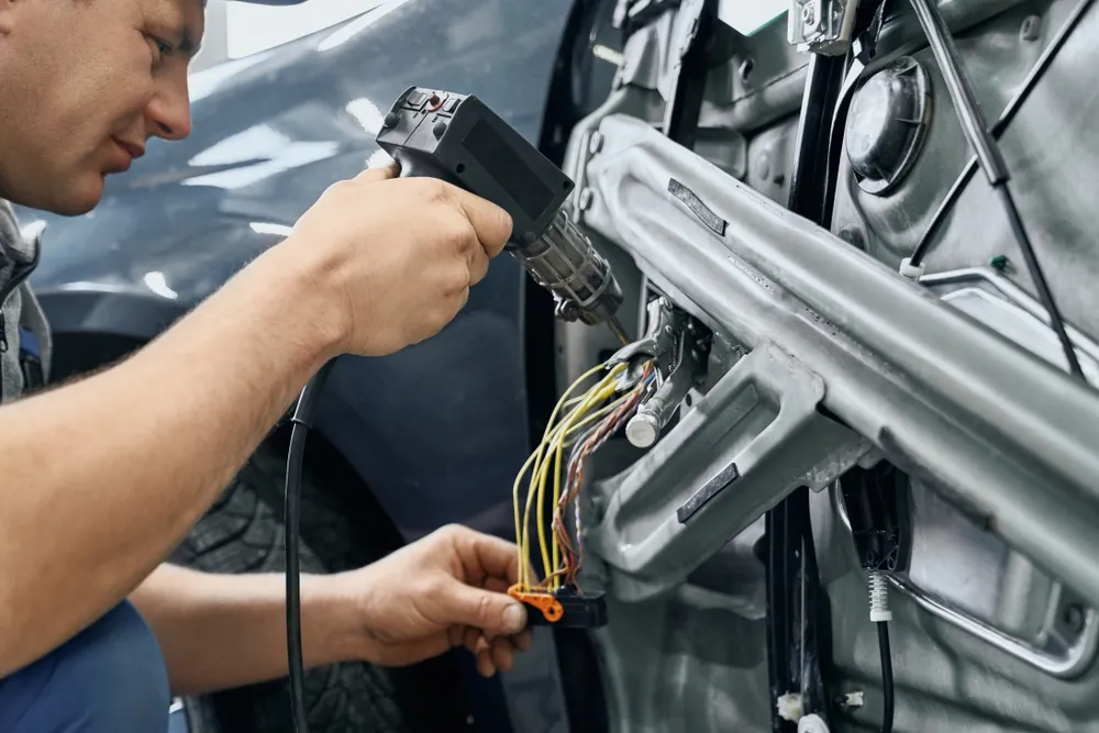 Side view of car electric repairman holding and using the soldering iron to blend the wires for repairing signaling in disassembled door of auto.