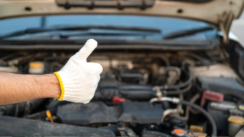 Action of a repairman in white cotton glove is thumbing up to confirm the good condition of the maintenance job with vehicle engine chamber as blurred background. Industrial working scene, close-up.